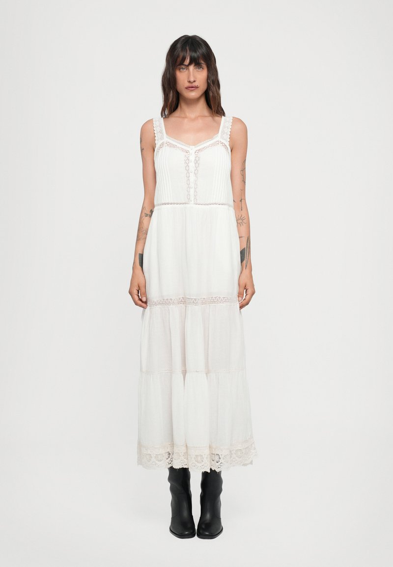 Vestido largo blanco con detalles de encaje, cuerpo plisado y falda en capas. Presenta tirantes ajustables y un delicado dobladillo de encaje. Se lleva con botas negras.