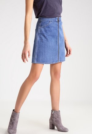 Personne portant une mini-jupe en denim bleu avec une fermeture éclair sur le devant, un haut sombre et des bottines en daim gris, debout devant un fond clair uni.