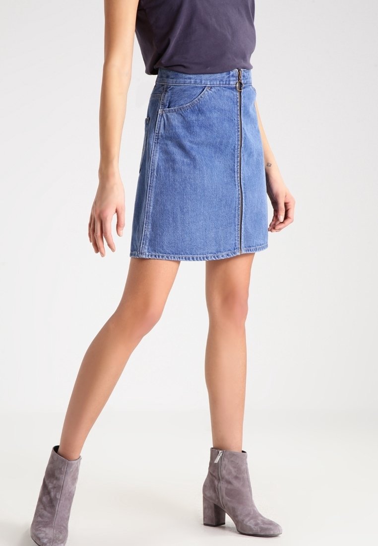 Personne portant une mini-jupe en denim bleu avec une fermeture éclair sur le devant, un haut sombre et des bottines en daim gris, debout devant un fond clair uni.