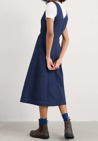 Džínové šaty s upnutým pasem, áčkovou sukní a funkčností. Mají hluboký V výstřih na zádech, kombinované s bílým žebrovaným tričkem a hnědými botami.