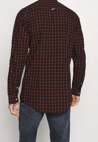 Karierte Bluse mit schwarzem Grund, rotem und orangefarbenem Gittermuster, Knopfmanschetten und abgerundetem Saum. Mit einer Schlaufe auf der Rückseite.