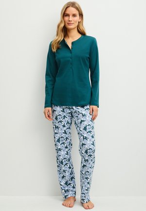 Femme portant un haut sarcelle à manches longues avec des boutons et un pantalon de pyjama blanc à motif floral bleu, debout pieds nus contre un mur blanc.