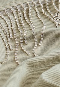 Bežinė medžiaga, puošta blizgančiais sidabro spalvos akmenukais, išdėstytais bangomis, kuriuos sudaro lašai, sukuriant tekstūruotą ir detalią puošybą.