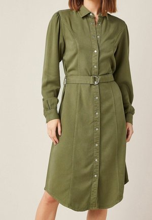 Kobieta ubrana w oliwkowozieloną sukienkę koszulową z długimi bufkowatymi rękawami i dopasowanym paskiem w talii.