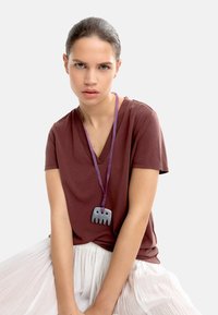 Brązowy t-shirt z dekoltem w serek w połączeniu z białą plisowaną spódnicą; nosi szary wisiorek w kształcie słonia na purpurowym sznurku.