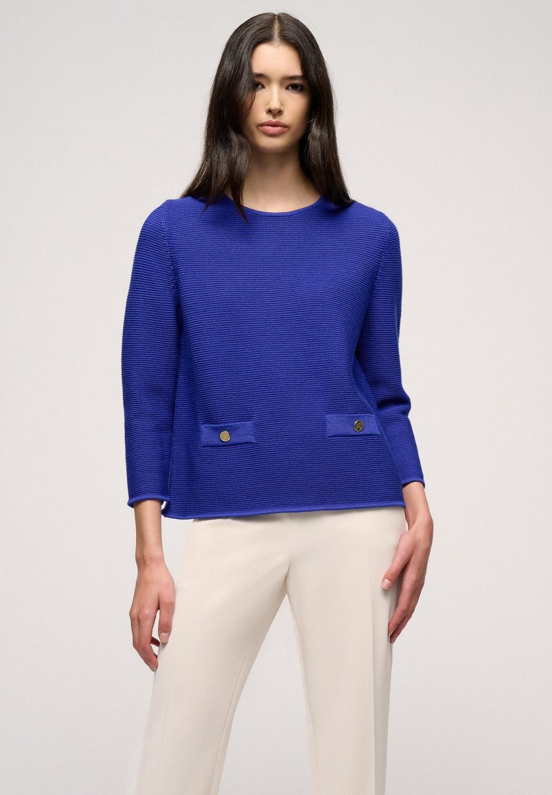 Heller blauer strukturierter Pullover mit langen Ärmeln, Rundhalsausschnitt und zwei Fronttaschen, die mit goldenen Knöpfen verziert sind. Kombiniert mit cremefarbenen Hosen.