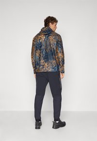 Veste à capuche de couleur bleu foncé avec un motif éclaboussé marron, arborant un design décontracté. Associée à un pantalon noir et des chaussures noires robustes.