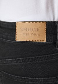 Jeans in denim nero con etichetta in pelle tan con testo in rilievo "2ND DAY THINKTWICE" sulla vita. Visibili dettagli delle cuciture.