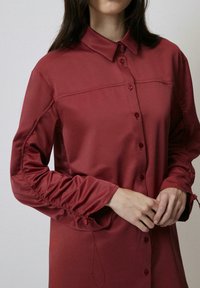 Camicia button-up bordeaux con una finitura in satin liscia. Presenta un colletto classico, tessuto arricciato sui polsini e una silhouette affusolata.