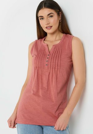 Vrouw met lang bruin haar, gekleed in een mouwloze top in stoffige rozentint met plooien en knopen aan de voorkant, gecombineerd met lichtblauwe spijkerbroek.