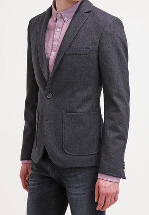 Homme portant un blazer gris foncé sur une chemise rose boutonnée et un jean foncé, debout devant un fond blanc uni.