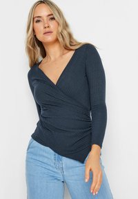 Dunkelblaues Langarmoberteil mit einem strukturierten Rippmuster, das ein V-Ausschnitt-Design und geraffte Details an der Taille aufweist. Kombiniert mit hellen Jeans.