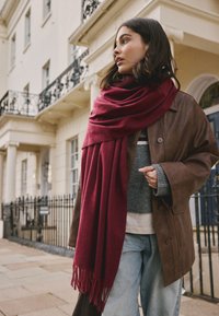 Bordeauxroter Schal mit Fransen, getragen über einem grauen Pullover und einer braunen Lederjacke. Lässige blaue Jeans vervollständigen das Outfit.