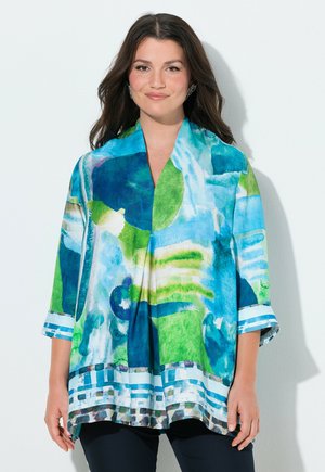 Kobieta ubrana w luźną bluzkę z długim rękawem z abstrakcyjnymi wzorami akwarelowymi w odcieniach niebieskiego, zielonego i białego, stojąca na jednolitym jasnym tle.