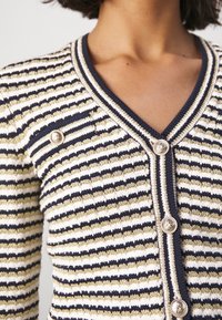 Kobieta w paskowanym granatowym, białym i złotym swetrze z dzianiny, z ozdobnymi złotymi guzikami i kołnierzem w serek.