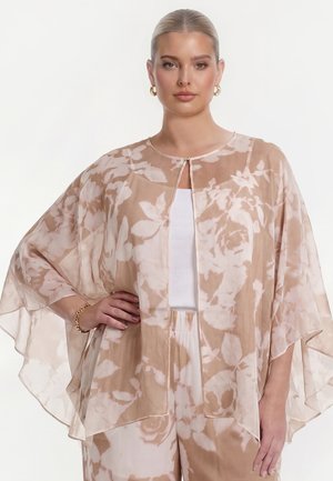 Vrouw met naar achteren gestyled haar, die gouden creolen oorbellen draagt, een doorschijnende beige bloemencape, een wit topje en bijpassende beige bloemenbroek, poserend met één hand op de heup.