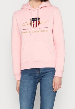 Personne portant un sweat à capuche rose clair avec le logo "GANT 1949 American Sportswear" et un jean bleu sur un fond clair uni.