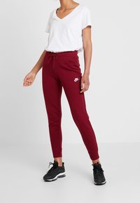 Burgundy joggers gjorda av mjukt tyg med elastisk midja och snörning. Vit Nike-logotyp på vänstra låret. Bärs med en vit V-ringad t-shirt.