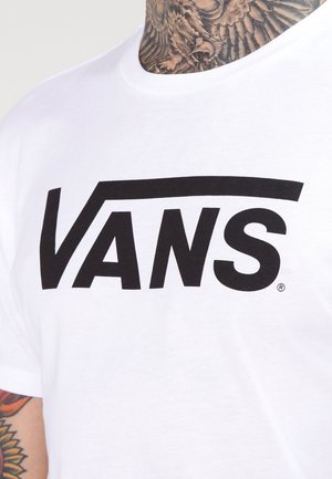 Biała koszulka z dużym czarnym logo Vans na piersi, noszona przez osobę z tatuażami pokazującą tatuaże na szyi i częściowo na ramieniu.