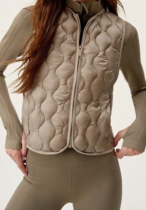 Nahaufnahme einer Person, die eine geschlossene, beige gesteppte Weste über einem langärmeligen, gerippten Oberteil und passenden Leggings trägt, die Hände auf den Hüften ruhend.