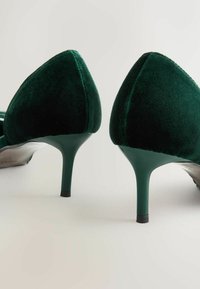 Et par mørkegrønne fløjl kitten heels med spidse tæer på en lys baggrund, vist fra bagsiden.