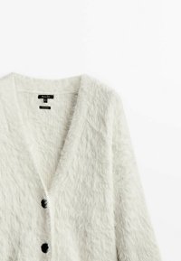 Cardigan bianco e soffice con scollo a V profondo, maniche lunghe e tre bottoni neri sul davanti, che mette in mostra un tessuto morbido e testurizzato.