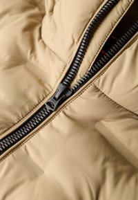 Tela de chaqueta acolchada beige con textura acolchada, con un zipper negro y una pestaña para tirar. La apertura del zipper está situada en el cuello.