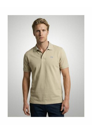 Blonde man die een beige korte mouwen polo draagt met een klein blauw logo op de linkerborst, staand voor een effen grijze achtergrond.