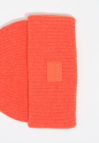 Gorro de punto naranja con un acabado texturizado y una etiqueta rectangular que muestra el nombre de la marca. Diseño plano, material suave, sin patrones visibles.