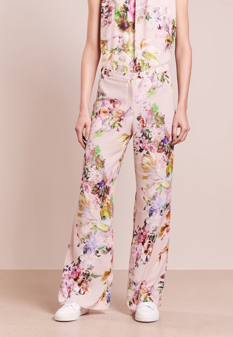 Bloemen jumpsuit met een lichtroze achtergrond; heeft levendige kleuren, brede benen en een getailleerde taille. Gecombineerd met witte sneakers.