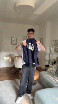 Ung mand står i en stue og holder en marineblå GAP-sweatshirt, iført en hvid t-shirt, sorte jeans og hvide sneakers.