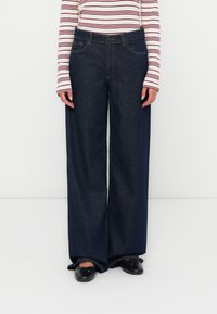 Jean en denim à jambes larges de couleur bleu foncé avec une texture lisse, dotés de poches avant et d'une taille classique. Porté avec un haut à rayures à manches longues.