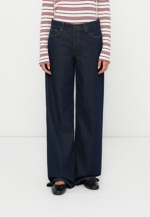 Jeans denim a gamba larga in blu scuro con una texture liscia, dotati di tasche frontali e una cintura classica. Indossati con una maglietta a righe a maniche lunghe.