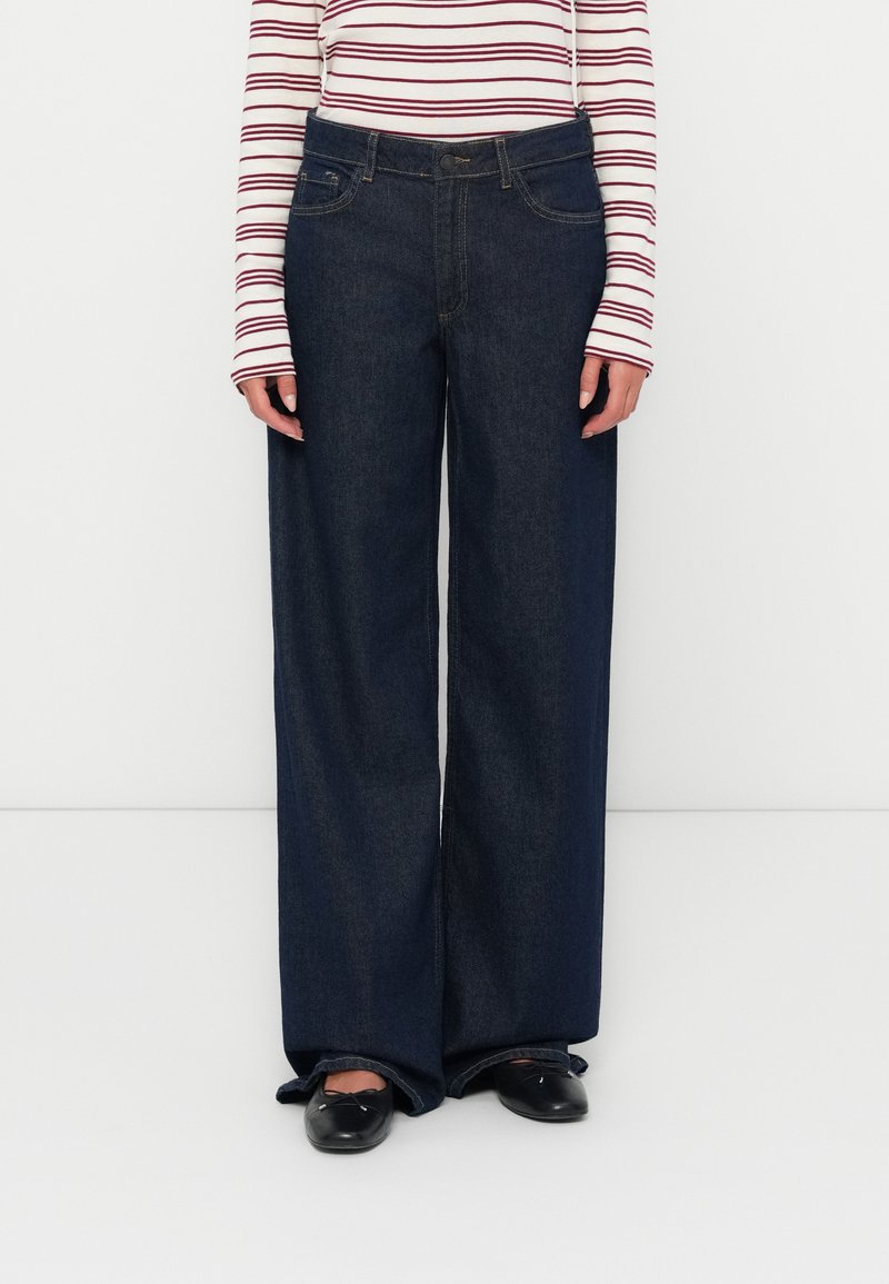 Jean en denim à jambes larges de couleur bleu foncé avec une texture lisse, dotés de poches avant et d'une taille classique. Porté avec un haut à rayures à manches longues.