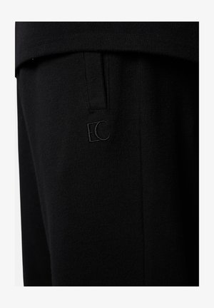 Schwarze Stoffhose mit einem dezenten "EC"-Logo in Stickerei nahe der Taschennaht, zeigt Strickstruktur und maßgeschneiderte Nähdetails.
