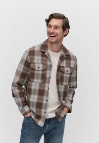 Brown and gray plaid shirt jacket with two front pockets, button closure, and a collared neck, worn over a light-colored t-shirt and jeans.