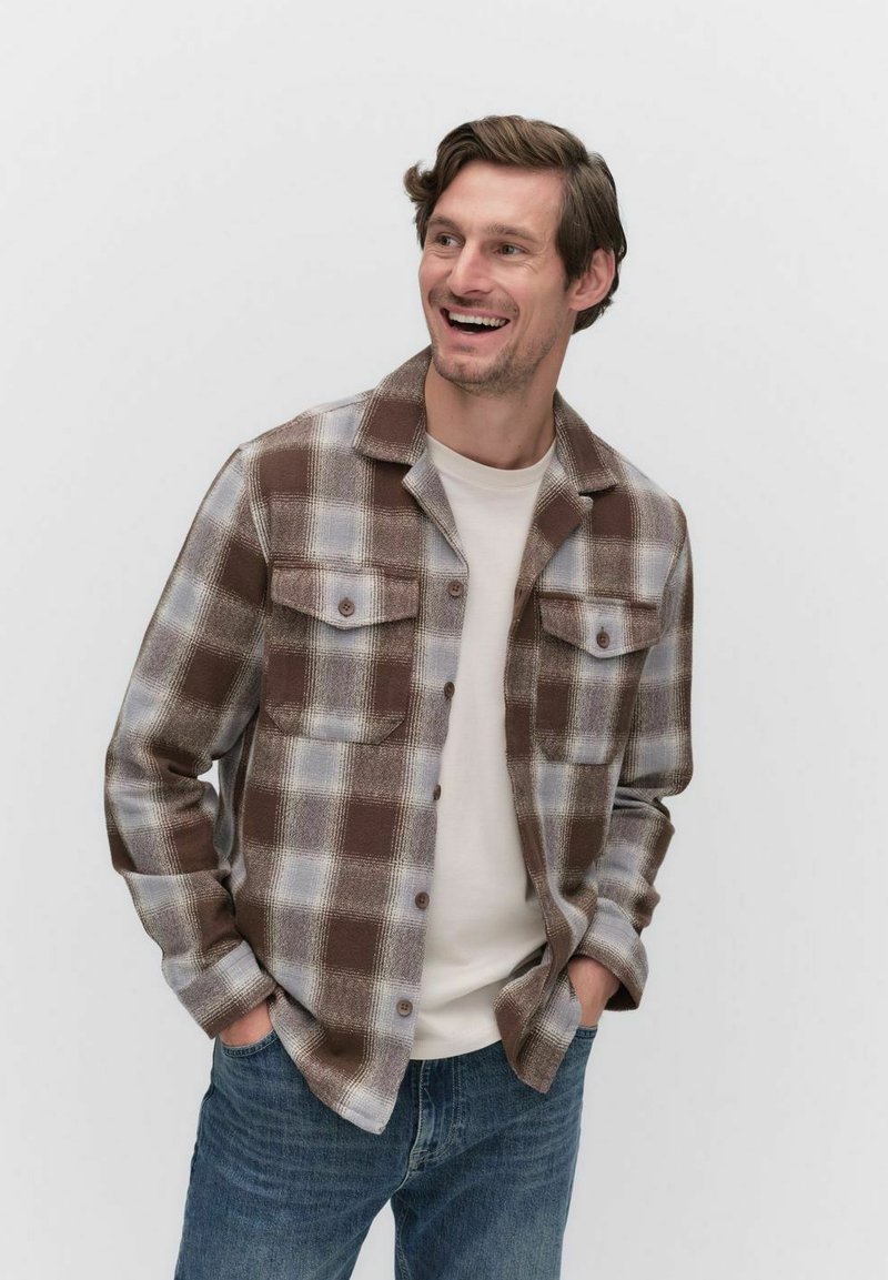 Brown and gray plaid shirt jacket with two front pockets, button closure, and a collared neck, worn over a light-colored t-shirt and jeans.
