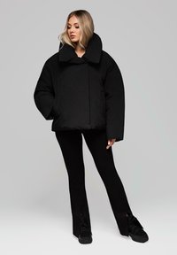 Černá péřová bunda s širokým límcem, oversized designem a dvěma bočními kapsami, doplněná o úzké černé kalhoty a černé tenisky.