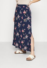 Jupe maxi florale bleu marine avec une fente sur le côté, fabriquée en tissu léger. Décorée de fleurs roses et blanches. Associée à des sandales blanches à lanières croisées.