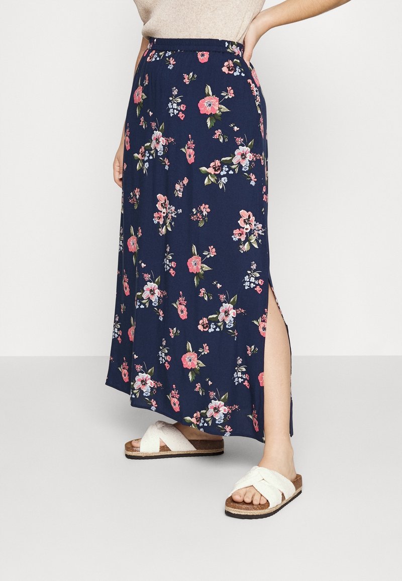 Jupe maxi florale bleu marine avec une fente sur le côté, fabriquée en tissu léger. Décorée de fleurs roses et blanches. Associée à des sandales blanches à lanières croisées.