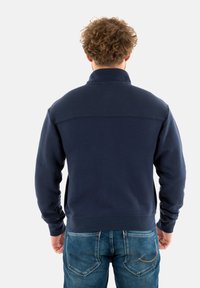 Felpa blu navy con colletto alto, texture liscia e polsi a coste, abbinata a jeans in denim chiaro con dettagli di cucitura delle tasche posteriori.