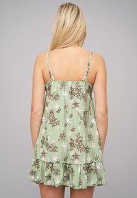 Rochie de vară cu imprimeu floral în verde deschis, cu flori maro și albe. Are bretele subțiri și un tiv evazat cu volane. Textură de țesătură moale.