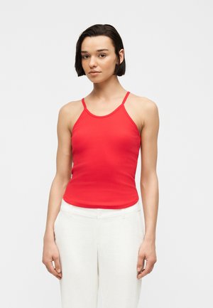 Jeune femme aux cheveux courts et foncés portant un top rouge sans manches et un pantalon blanc, debout devant un fond clair uni.