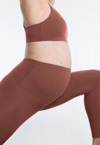 Personne enceinte en leggings et brassière de sport couleur rouille réalisant une fente de yoga sur un fond uni.