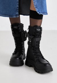 Bottes de combat noires avec une semelle robuste, un laçage à l'avant, une fermeture éclair sur le côté et des détails de sangle à boucle réglable. Fabriquées en cuir synthétique.