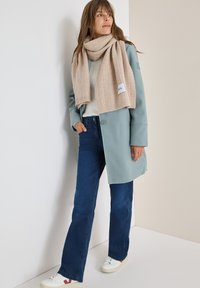 Mujer vestida con abrigo azul claro, bufanda de punto beige, suéter blanco, jeans azul oscuro y zapatillas blancas, de pie frente a una pared blanca y beige.