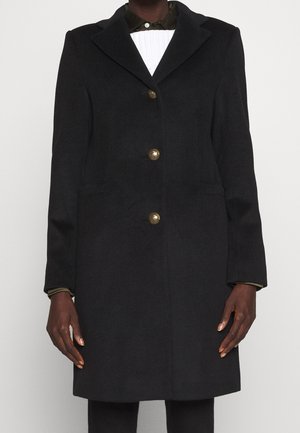 Manteau noir simple boutonnage avec trois boutons dorés porté sur une chemise blanche avec col et poignets visibles.