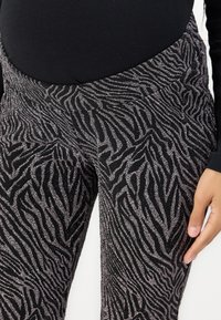 Svarte leggings med et sølvfarget zebra-stripe mønster, med en glatt tekstur og en komfortabel midjebånd. Hånd synlig på siden.