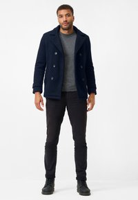 Marinblå sjöortsjacka med stora knappar och bred krage, parat med en grå tröja och svarta slim-fit byxor. Modell har på sig svarta snörstövlar.