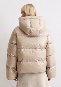 Beżowa pikowana kurtka puchowa z kapturem, z przeszywanymi panelami i błyszczącą fakturą. Ma luźny krój i szerokie rękawy.